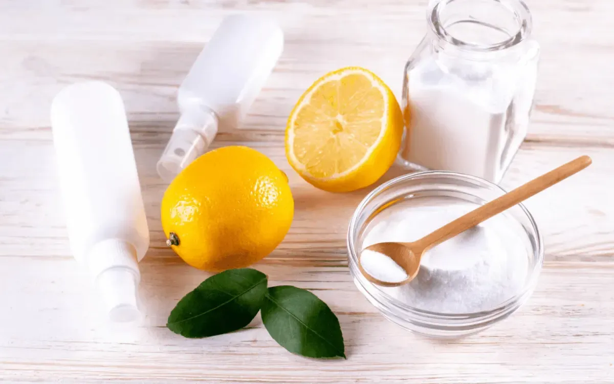 A flat lay of natural cleaning supplies including lemons, baking soda in glass containers, spray bottles, and green leaves on a wooden surface.