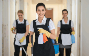 Group of professional cleaners in uniforms and aprons smiling while carrying cleaning supplies and mops in a hallway.