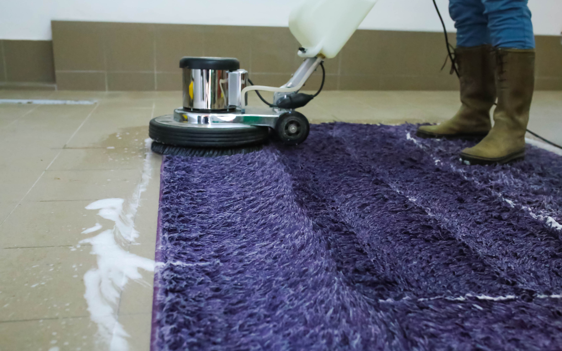 Worker cleaning a purple carpet with a professional floor scrubbing machine and soap foam on tiles.